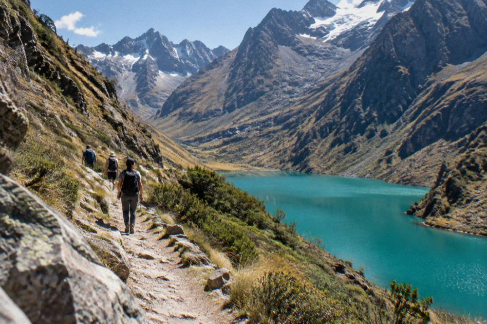Hikers on a mountain path above a turquoise glacial lake surrounded by rocky peaks