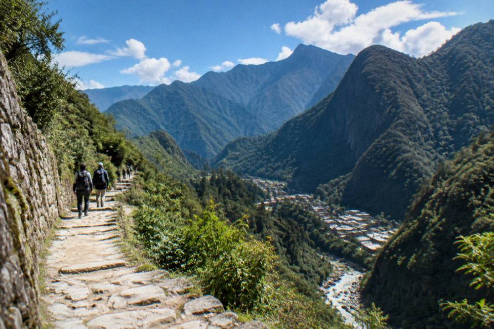A stone terrace path above a green valley with hikers walking in the distance