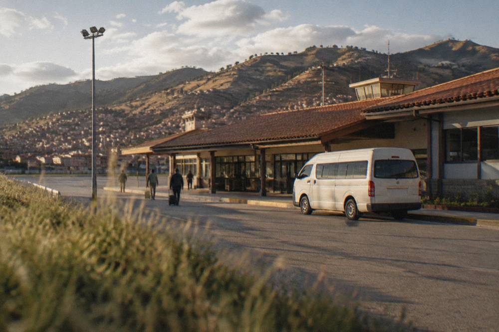 A parked white van at a kerb outside a low airport building with hills behind it