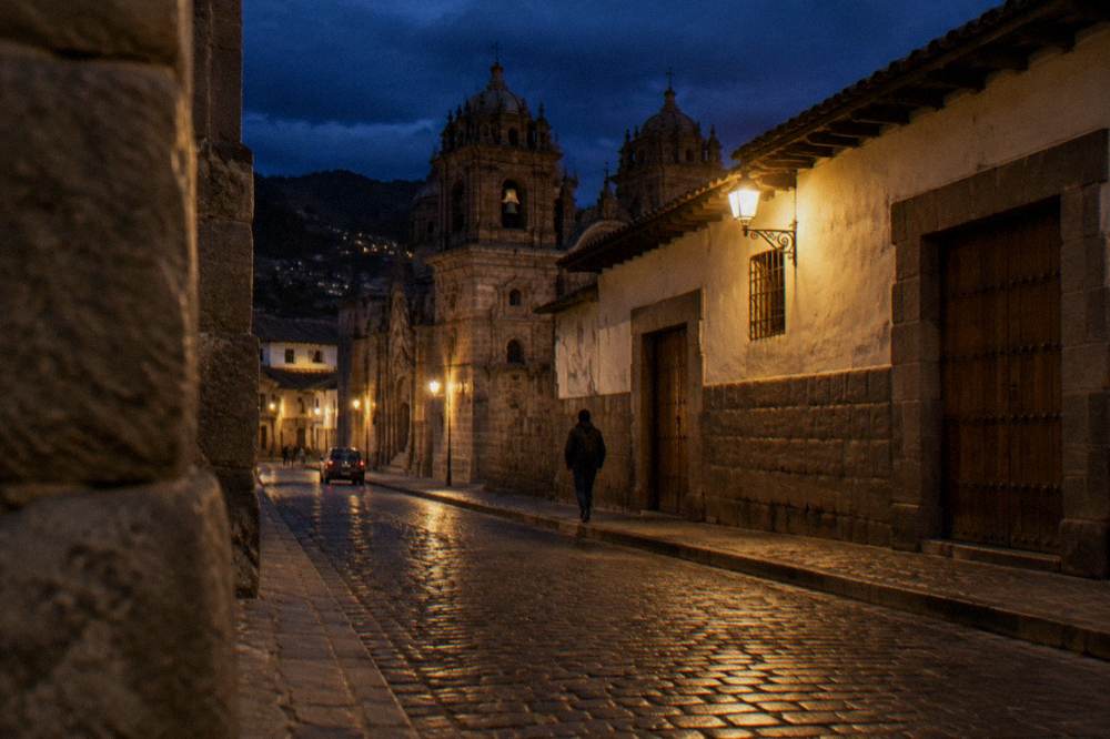 A lit stone street in Cusco with closed wooden doors and a person walking under a streetlamp