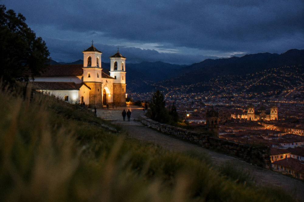 The lit towers and tiled roofs of Cusco seen from a dark hillside viewpoint at night