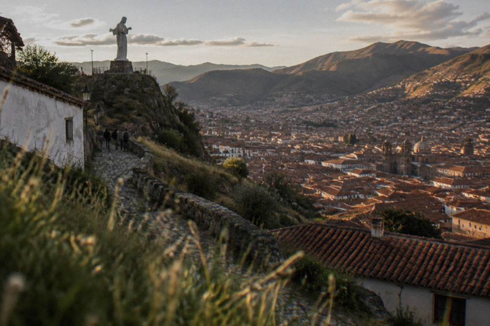 A white statue on a hilltop overlooking a dense red-roofed city in a mountain basin