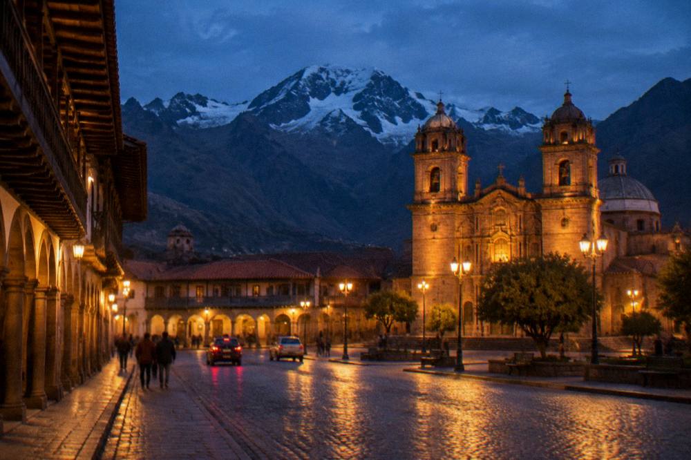 A city square at dusk with lit church facades and colonnaded buildings under a deep blue sky
