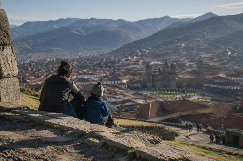 A parent and child resting on a stone step overlooking red rooftops of Cusco