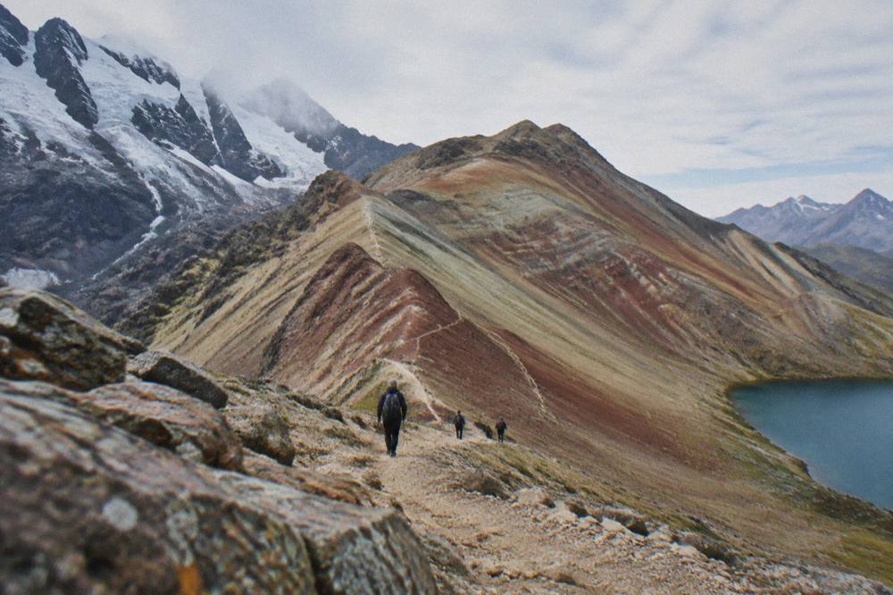 Hikers descending a red and ochre striped mountain ridge under a bright high-altitude sky