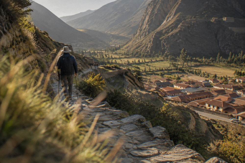 A hiker in a warm jacket and sun hat walking a stone Inca path above a valley