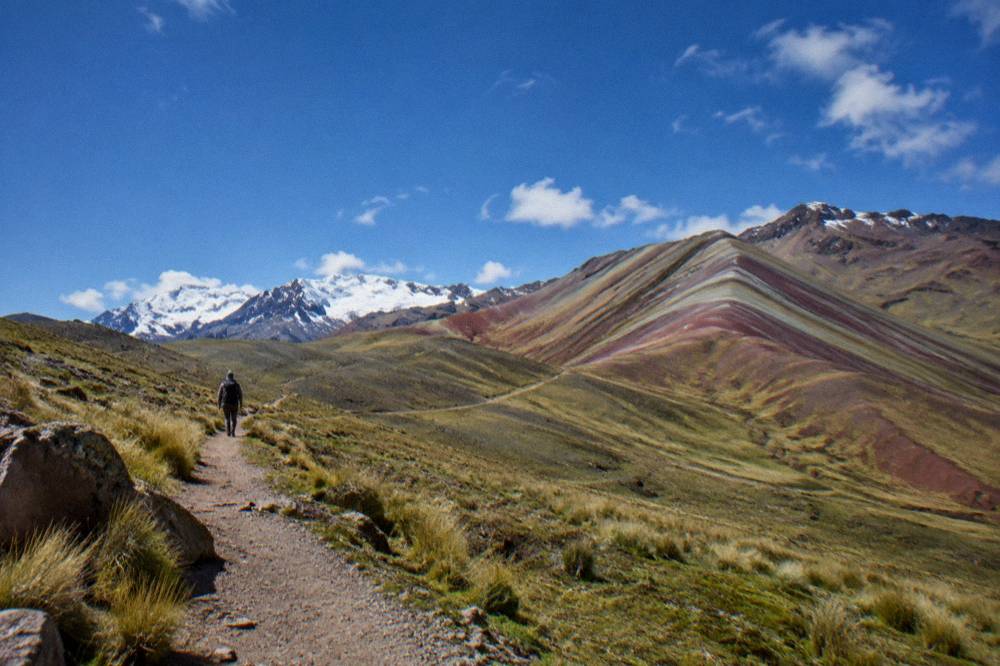 Rolling high Andean grassland with distant snow peaks and a small hiker on a dirt path
