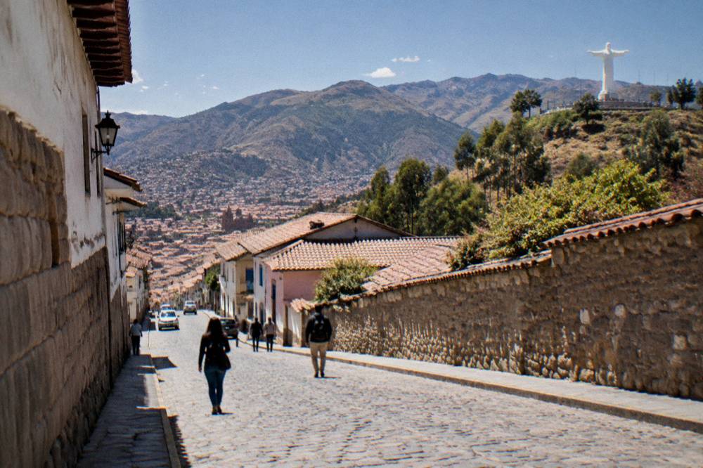 Visitors walking a sunlit cobblestone street in central Cusco with mountains in the distance