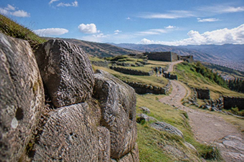 Rolling grass hills above Cusco with stone terrace walls and a dirt path leading to a ruin site