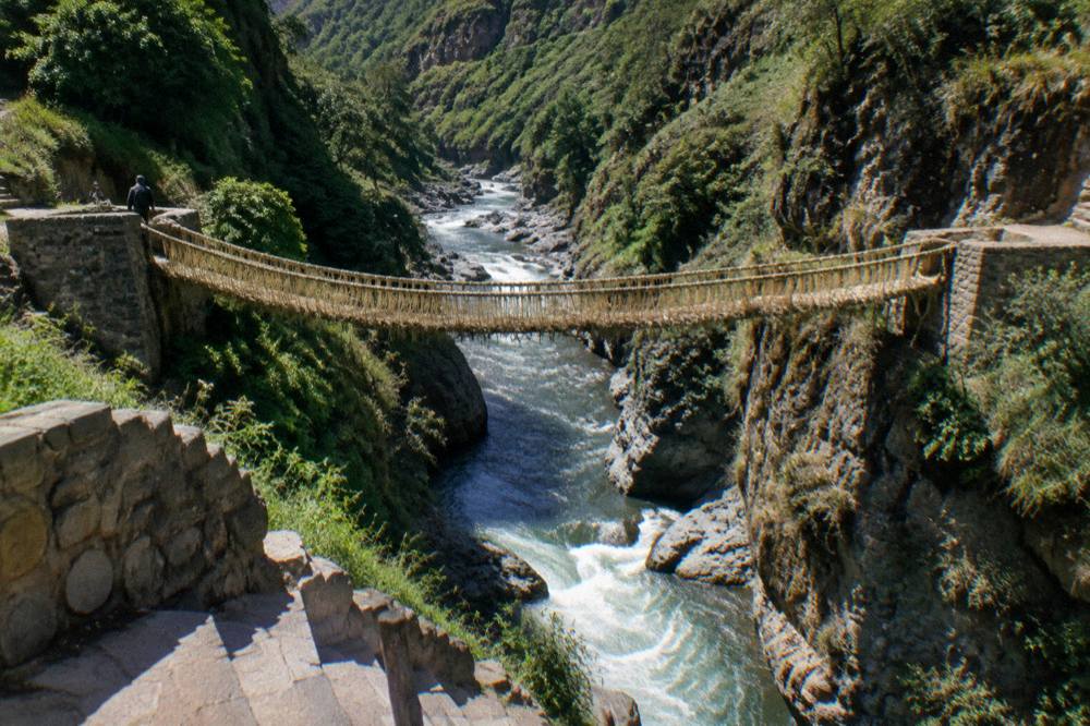 A woven grass bridge spanning a deep river gorge between green canyon walls