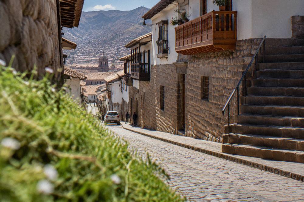 A narrow cobbled street in the Cusco historic centre with stone walls, wooden balconies and a stone staircase