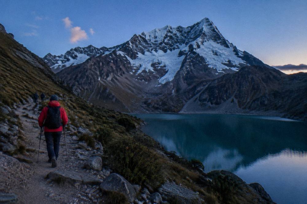 Trekker in a red jacket walking a high Andean path under a clear blue sky