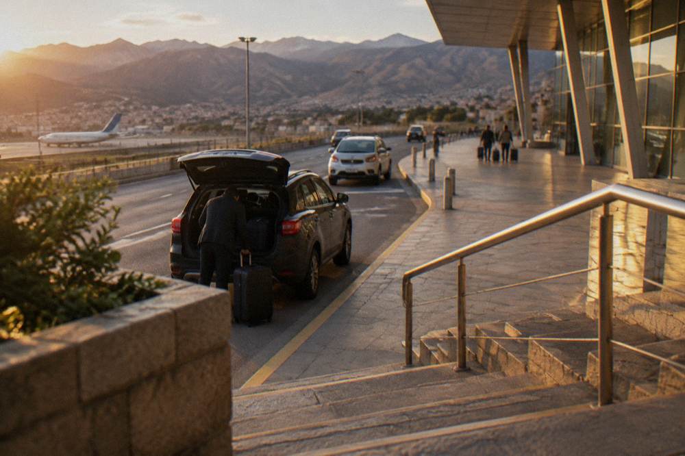 A driver loading suitcases into the boot of a car outside an airport terminal