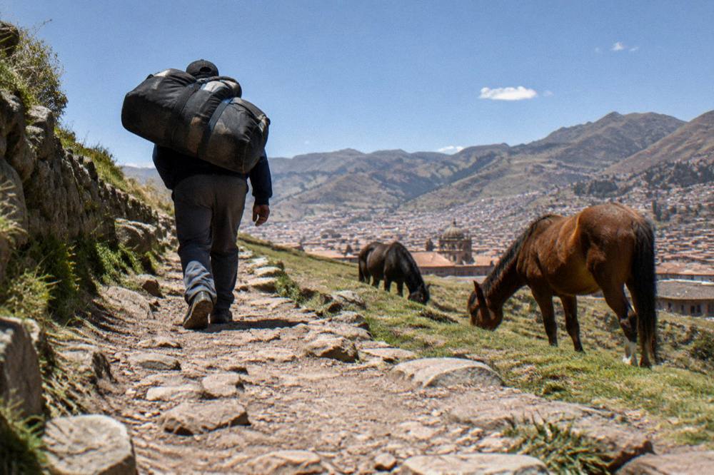 A porter with a loaded duffel bag walking a mountain trail past grazing horses