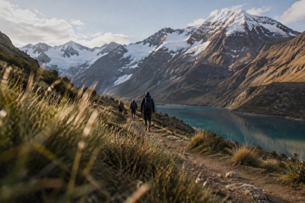 Hikers with backpacks walking a dirt path through high grassland with distant snowy mountains