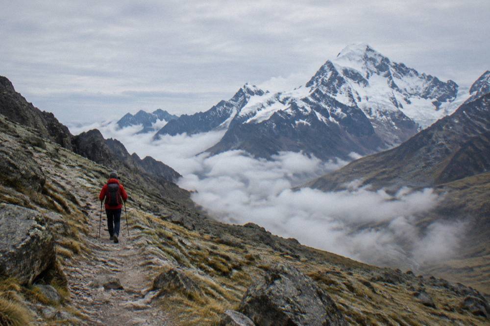 A trekker in a red jacket crossing a high mountain pass with clouds in the valley below