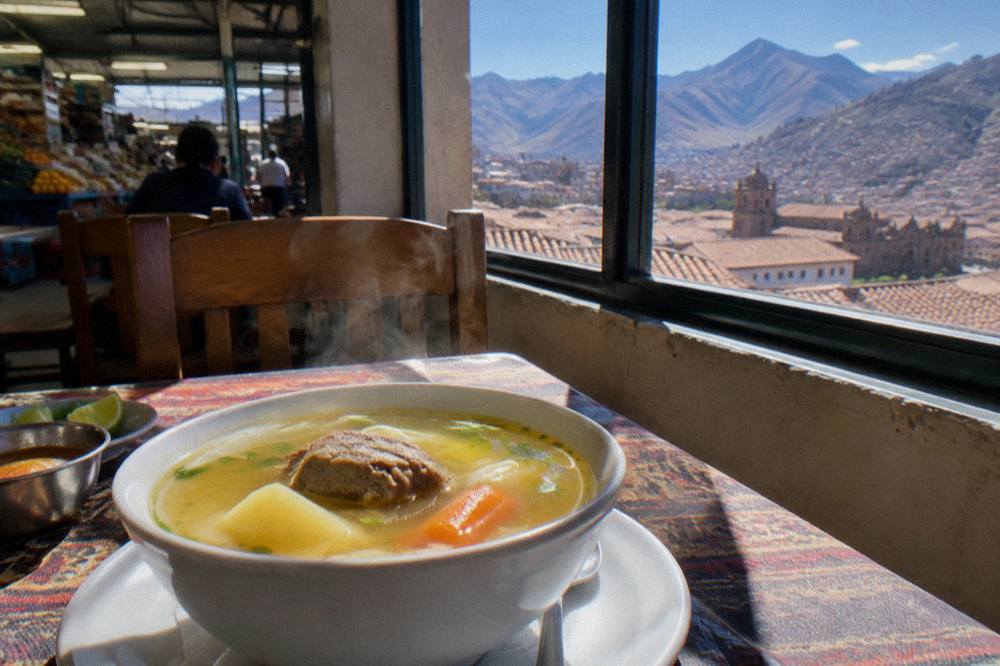A steaming bowl of Andean soup on a table with mountains visible through a window