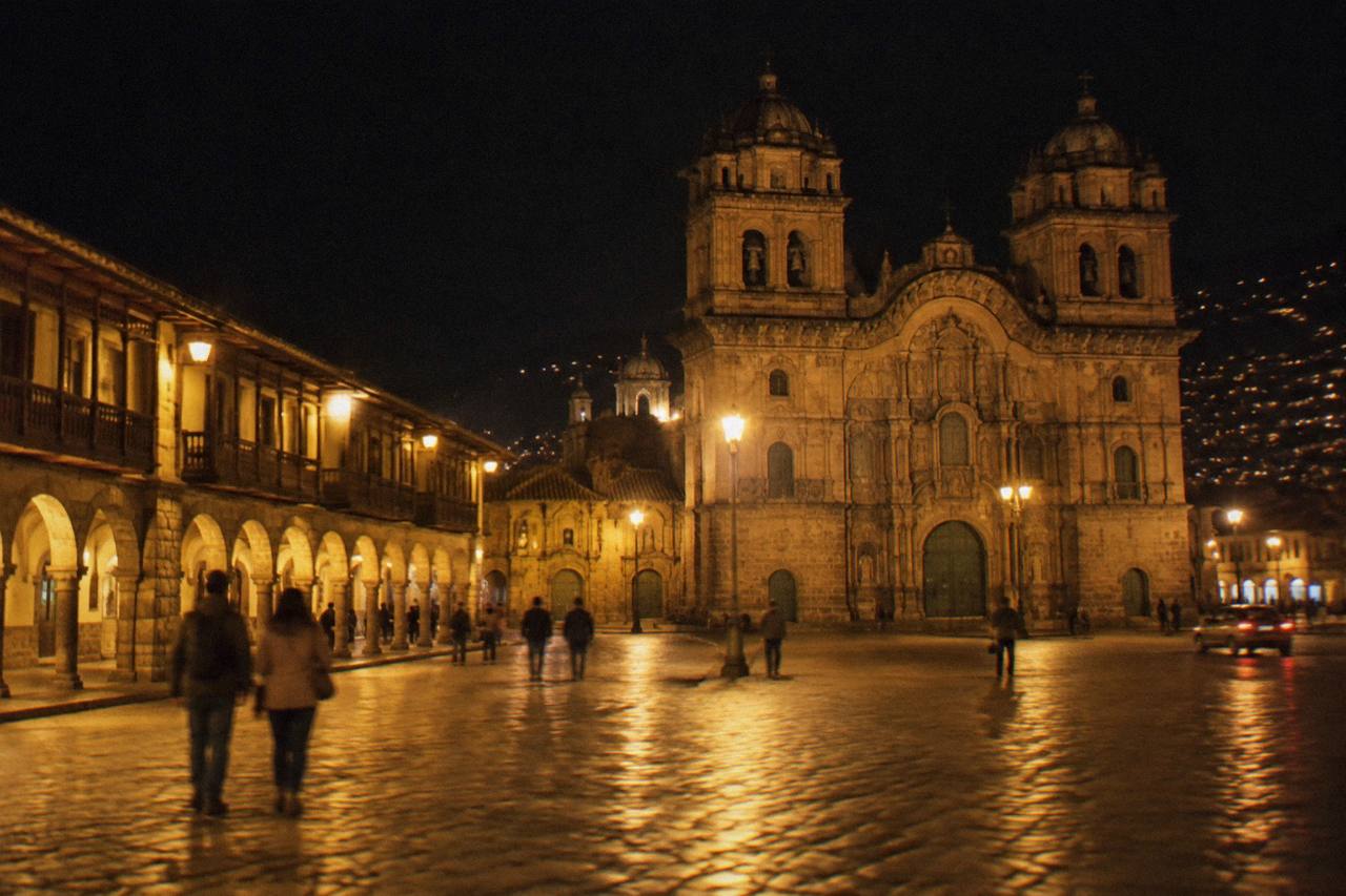 The Plaza de Armas of Cusco after dark, the arcades and cathedral facade lit warmly, a few people crossing the stone-paved square