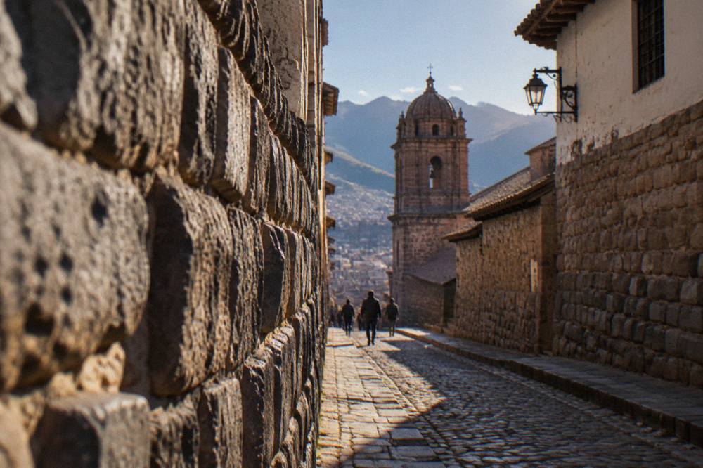 Narrow stone-walled lane in Cusco rising toward a church tower in morning light