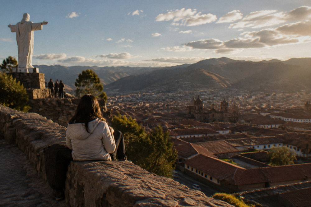 A visitor in a light jacket resting on a stone wall above red-tiled roofs of a high Andean city