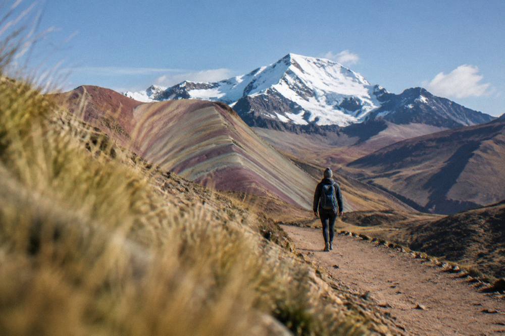 A hiker with a small backpack walking a dirt trail toward a snow-capped Andean peak